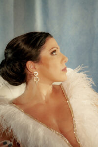 Woman in jeweled-trimmed white feathered dress and sparkling earrings, dark hair in low bun, poses against soft blue background.