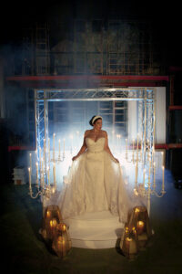 Bride in white gown on lit platform with candelabras and lanterns, misty backdrop—timeless Gulf South wedding inspiration.