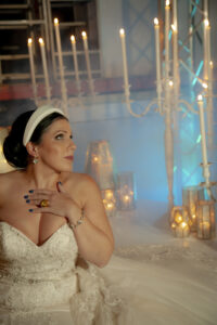 Bride in white dress sits by lit candles, hand on chest, with dreamy warm and blue lighting for romantic Louisiana wedding.