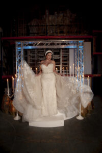 Bride in strapless ornate gown on white platform, holding her train, surrounded by candles; elegant Gulf South wedding scene.