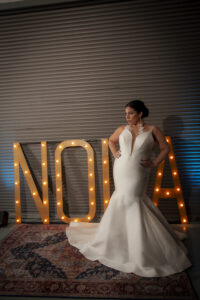 Bride in white gown stands on patterned rug before large NOLA marquee letters against corrugated metal at Louisiana wedding.