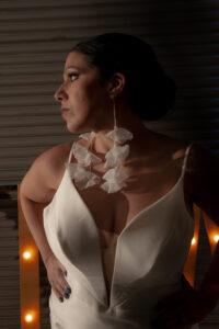 Bride in a white dress with ruffled earrings, hair pulled back, stands in dramatic light with round lights behind her.