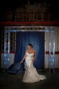 Woman in white strapless gown by blue chair, metal frame and dark blue curtain, evoking New Orleans wedding elegance.