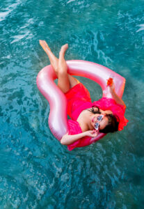 Woman in pink swimsuit and sunglasses lounges on a pink pool float with a drink, surrounded by clear blue water.