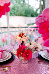 Pink sequin table with elegant plates, gold utensils, red and white floral centerpiece, and pink balloons outdoors.