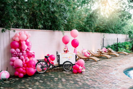 Pink ice cream cart with balloons by a pool, lounge chairs, and greenery creates a festive outdoor party scene.