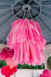 Pink and light pink paper streamer chandelier hangs from a wooden gazebo, with pink balloon clusters in the background.