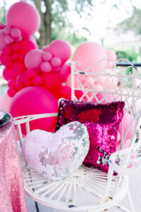 White ornate chair with silver heart-shaped and pink sequin pillows beside bright pink balloons and a pink sequin table.