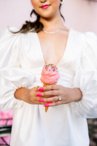 Woman in white dress holding pink ice cream with rainbow sprinkles, pink nails, jewelry, and a soft pink blurred background.