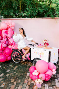 Woman in white dress sits on decorated ice cream bike cart with balloons and flowers against a pastel pink, festive backdrop.