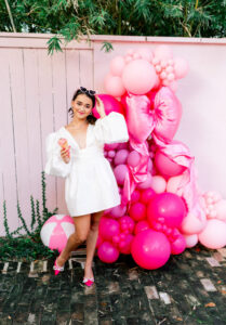 Woman in white dress holding ice cream cone by pink balloon display, pink fence, and greenery; wearing pink shoes, sunglasses on head.