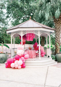 White gazebo with pink and white balloons, pink fringe canopy, and sequin tablecloth in a lush outdoor setting with palm trees.