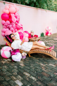 Woman in white dress relaxes on wooden chair with pink balloons, flowers, and beach balls against a pink wall on brick patio.