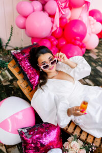 Smiling woman in a white dress relaxes on a chair with pink sequin pillows, holding a drink, surrounded by pink balloons and a beach ball.