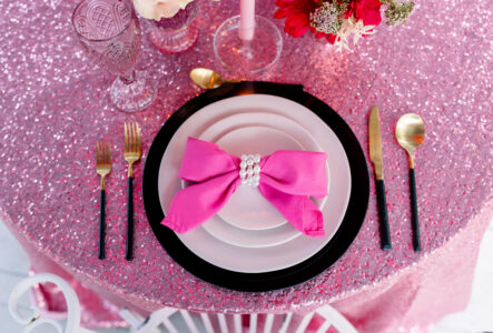 Chic table with pink sequin cloth, white and black plates, pink napkin with pearl ring, gold cutlery, flowers, and candle.