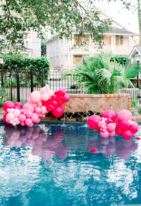 Clusters of pink, red, and white balloons float by a pool, with palm trees, a fence, and houses in the background.