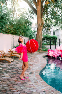 Woman in a bright pink dress and heels holds a large red heart by a pool with pink balloon decorations.