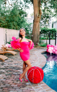 Woman in pink dress and heels poses by pool with red flowers, pink balloons, benches, trees, and brick patio.