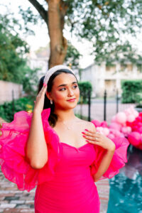 Woman in a bright pink ruffled dress by a pool with pink balloons, smiling and touching her headband; trees and houses behind.
