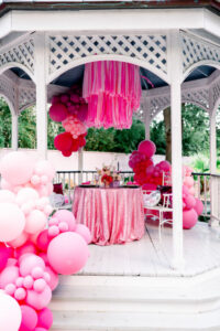 White gazebo with pink balloons, a pink tablecloth, fringe, and a table for two. Green trees in the background.