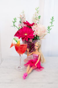Barbie doll in pink dress next to a pink watermelon cocktail and vase of pink and white flowers on marble surface.