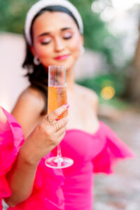 Woman in bright pink dress and pearl headband smiles softly, holding champagne with a blurred outdoor background.