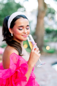 Woman in a pink ruffled dress and pearl headband smiles with closed eyes, holding champagne outside with greenery behind her.
