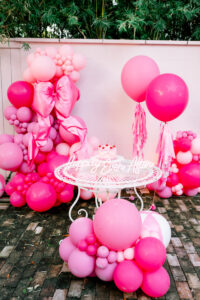 Outdoor white table with cake, Happily Ever After sign, pink and red balloons, bow, white fence and greenery backdrop.