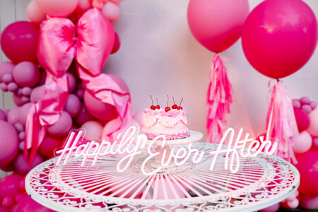 Pink cake with cherries on a white table, “Happily Ever After” sign, pink balloons, and festive decorations.