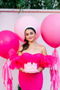 Woman in bright pink dress holding small pink cake, standing by large pink balloons against pale pink wall with greenery above.