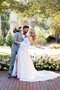 Groom in gray suit dips and kisses bride in white gown outdoors, surrounded by greenery and burgundy-white florals.