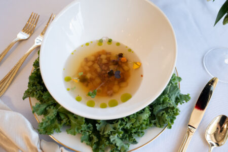 Clear broth with diced vegetables and herbs in a white bowl, plated with kale and gold utensils on a white tablecloth.