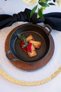 Grilled meat with red sauce and sage in a cast iron pan on a wooden board, served on a gold-rimmed plate.