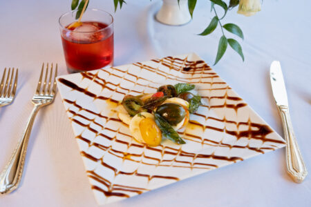 Caprese salad on a square plate with balsamic drizzle, fork and knife, red cocktail, greenery, and white tablecloth.