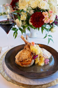 Grilled pork chops with fruit compote on a dark plate, served beside red wine and a floral centerpiece.