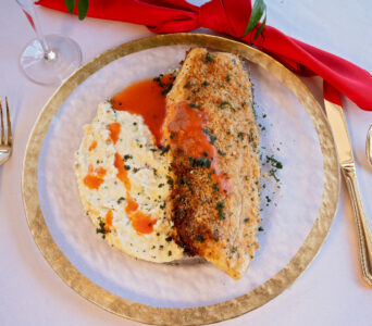 Breaded fish fillet with red sauce and mashed potatoes, garnished with herbs, on a white plate with gold rim.