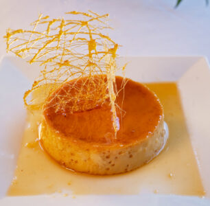 Creamy round flan on a white plate, topped with caramel syrup and sugar decoration, with sauce pooled around it.
