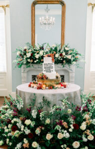 White wedding cake with fruit on a gray-covered table, surrounded by greenery and flowers by a decorated fireplace and mirror.