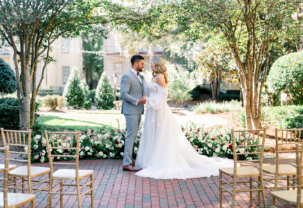 Bride and groom stand in a sunlit garden with flowers and gold chairs, wedding attire highlighted by sunlight.