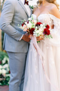 Bride in white gown with red and white bouquet stands by groom in light gray suit; faces not shown.