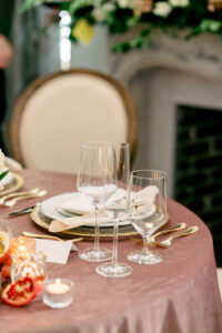 Stylish table with pink cloth, gold-rimmed plates, white napkins, three wine glasses, gold cutlery, candles, and fruit.