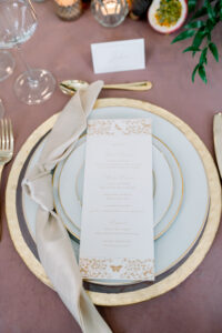 Gold-rimmed plates, beige napkin, menu card, and gold cutlery on a mauve tablecloth with candles and greenery.