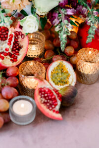 Pomegranate halves, grapes, passion fruit, figs, and glass candles on a table with flowers in the background.