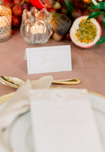 Elegant table setting with gold-rimmed plate, menu, gold cutlery, “John” name card, candles, figs, grapes, and passion fruit.