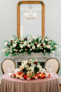 Round table with mauve tablecloth, floral centerpiece, pomegranates, candles, two chairs, and gold mirror in background.