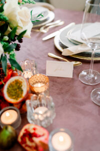 Bright table with mauve cloth, candles, flowers, fruits, plates, cutlery, wine glass, and John name card in front.