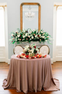 Round table with mauve cloth set for two, floral centerpiece, pomegranates; mantel, bouquet, gold mirror reflecting chandelier.