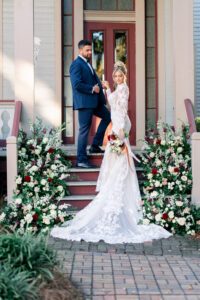 Bride in lace gown with bouquet stands on outdoor steps, looking back at groom; flowers decorate both sides of stairs.