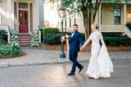 Bride in lace gown and groom in navy suit walk hand in hand on a brick street, smiling by a decorated house with greenery.