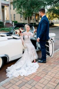 Photo: Sarah Alleman Photography Bride in lace dress in white vintage car holds bouquet as groom in navy suit helps her out; trees and houses in background.
